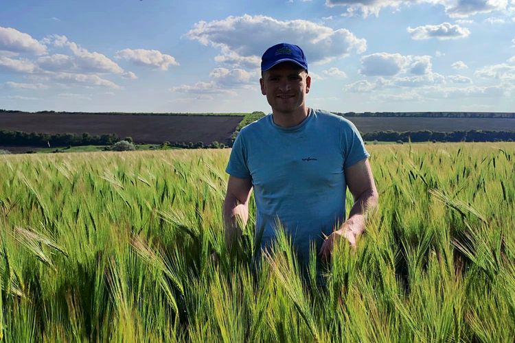Ein Mann steht lächelnd in einem grünen Feld mit reifendem Getreide unter einem blauen Himmel mit weißen Wolken. Er trägt ein hellblaues T-Shirt und eine blaue Kappe. Im Hintergrund sind weitere Felder und Bäume zu sehen.