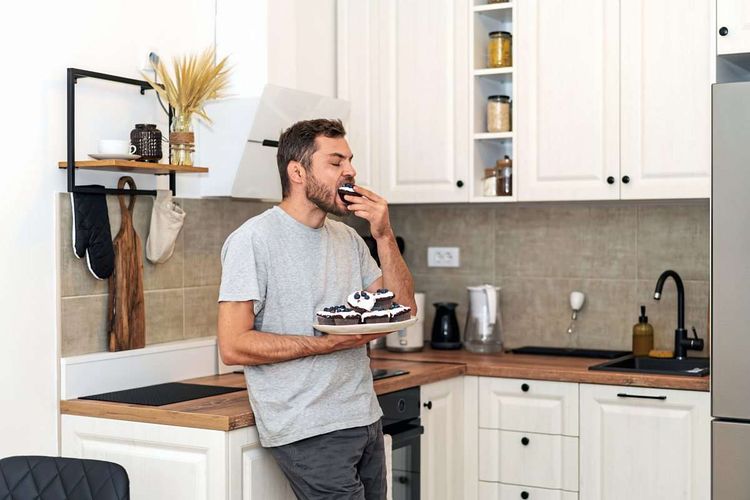 Junger Mann steht in der Küche und isst einen Muffin. Er hält in der Hand Teller mit weiteren Muffins