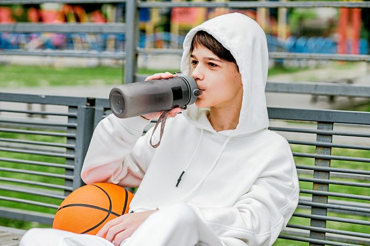 Ein Jugendlicher sitzt mit Hoodie und Basketball auf einer Bank und trinkt aus einer Trinkflasche