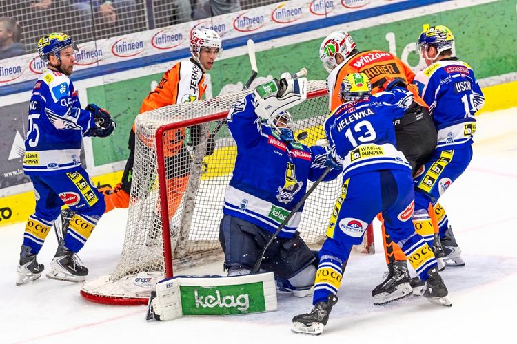 Eishockeyspielszene: Der Torwart des EC VSV, Joe Cannata, kniet vor dem Tor und hebt seinen Handschuh, während mehrere Spieler beider Teams, darunter Mark Katic und Adam Helewka vom EC VSV sowie Paul Huber von den Graz 99ers, um den Puck kämpfen. Das Geschehen findet während eines Spiels der win2day ICE Hockey League statt. Im Hintergrund sind die Bande und Zuschauer sichtbar.