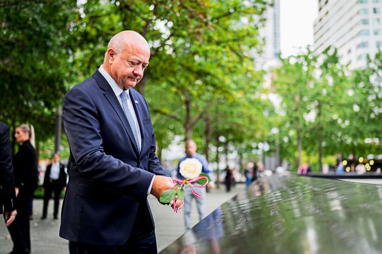 Ein Mann in Anzug legt eine Blume am 9/11-Memorial in New York City nieder. Im Hintergrund sind Bäume und unscharfe Passanten zu sehen.