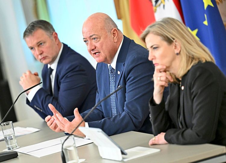 Drei Personen sitzen bei einer Pressekonferenz oder Sitzung an einem Tisch mit Mikrofonen. Im Hintergrund sind die Flaggen der EU und Österreichs zu sehen. Dokumente, ein Tablet und ein Glas Wasser befinden sich auf dem Tisch.