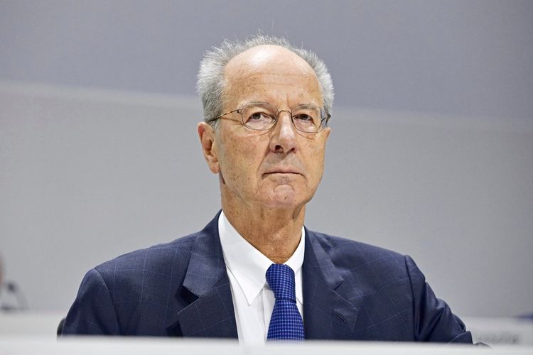 Ein älterer Mann mit Brille und grauem Haar trägt einen dunklen Anzug, ein weißes Hemd und eine blaue Krawatte. Er sitzt und blickt ernst in die Kamera. Der Hintergrund ist neutral gehalten.