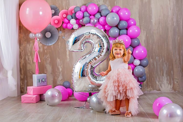 Kleines Mädchen in einem Prinzessinnenkostüm steht vor einer Fotowand mit pinken Luftballons.