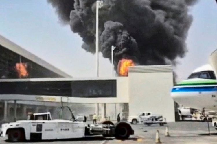 Schwarzer Rauch und Flammen steigen aus einem Gebäude am King Khalid Flughafen in Riad, Saudi-Arabien, auf. Im Vordergrund sind das Flughafengelände, Bodenfahrzeuge und Teile eines Flugzeugs sichtbar.
