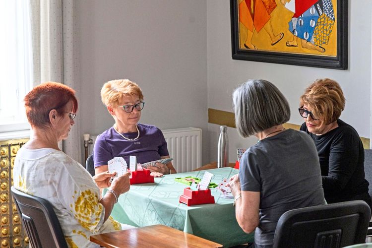 Vier ältere Frauen sitzen an einem Tisch in einem Raum und spielen Bridge. Auf dem Tisch liegen Karten und ein Bietkasten. Im Hintergrund ist ein buntes Gemälde an der Wand zu sehen.
