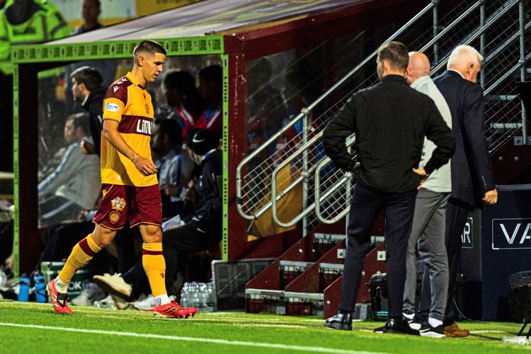 Ein Fußballspieler von Motherwell, gekleidet in einem gelb-roten Trikot, verlässt das Spielfeld nahe der Trainerbank. Im Hintergrund sind weitere Personen, darunter Trainer und Betreuer, sowie ein Schild mit 
