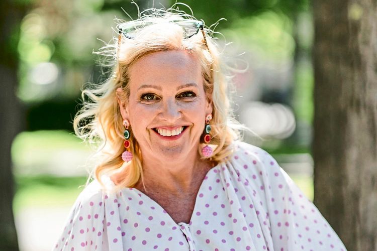 Eine Frau mit blonden Haaren steht lächelnd im Freien. Sie trägt eine weiße Bluse mit rosa Punkten, blaue Jeans, bunte Ohrringe und eine Armbanduhr. Im Hintergrund sind Bäume und ein unscharfer Park zu sehen.