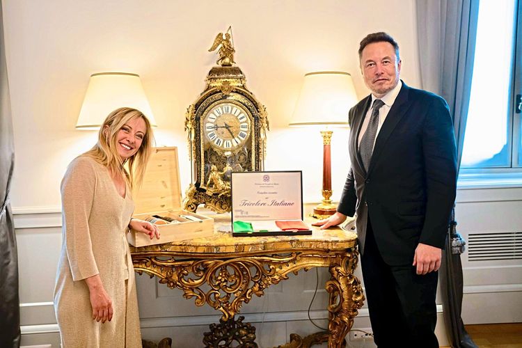 Die britische Premierministerin Giorgia Meloni und Tesla-Chef Elon Musk posieren gemeinsam für ein Foto. Zwischen ihnen steht ein goldener Tisch, auf dem unter anderem die italienische Flagge präsentiert wird.