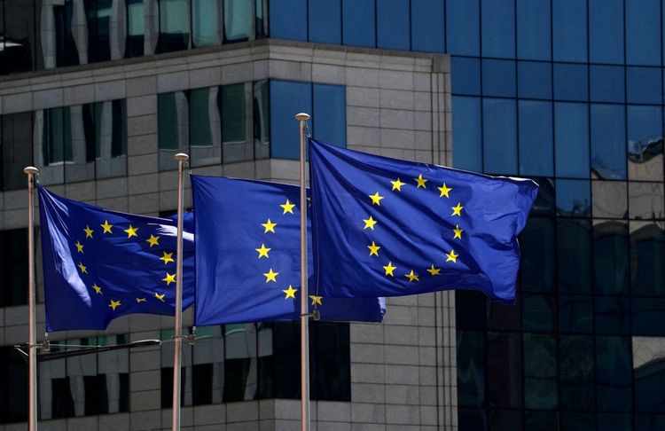 Drei Flaggen der Europäischen Union wehen vor dem Hauptsitz der Europäischen Kommission in Brüssel, Belgien. Im Hintergrund ist ein modernes Bürogebäude mit Glas- und Steinfassade zu sehen.