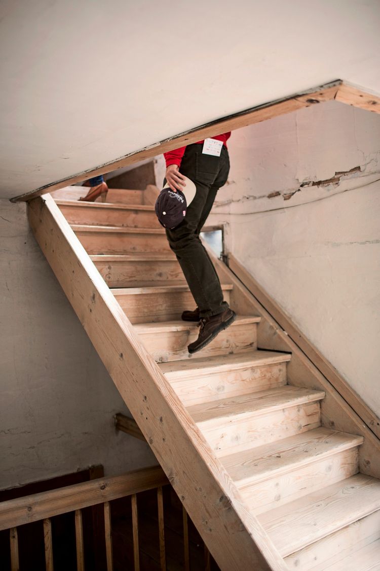 Eine Person geht eine hölzerne Treppe in einem Innenraum hinauf. Die Wand neben der Treppe zeigt Spuren von Abnutzung. Die Szene deutet auf eine Renovierung oder Umbauarbeiten hin.
