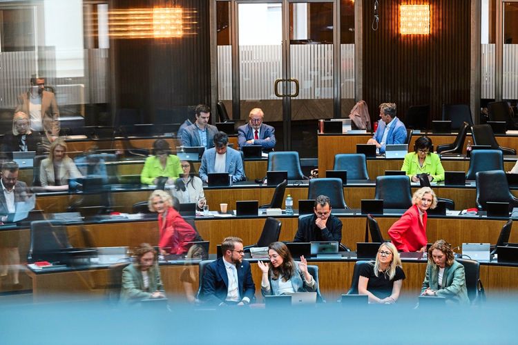 Eine Szene aus einem Parlamentssaal mit Abgeordneten an ihren Plätzen. Personen arbeiten an Laptops, diskutieren oder schauen in Unterlagen. Im Hintergrund und Vordergrund sind verschiedene Personen in Gespräch vertieft.