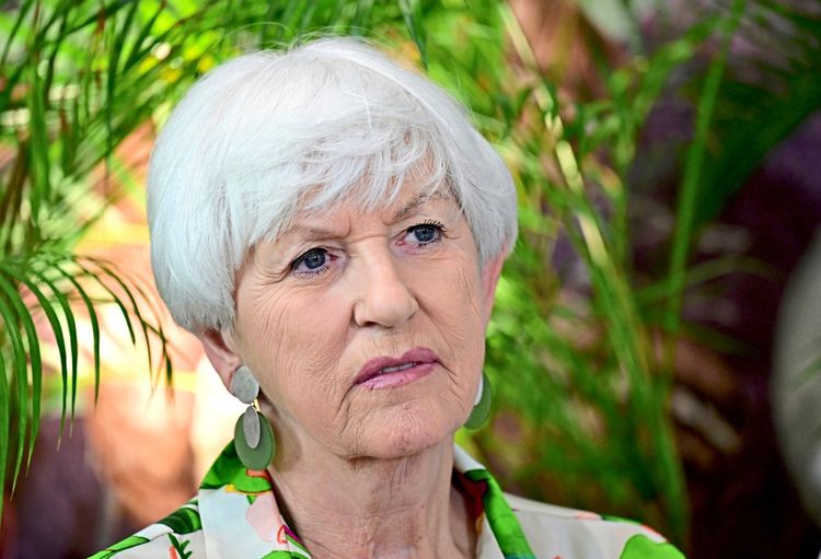 Eine ältere Frau mit weißem Haar und großer Ohrringe blickt ernst. Im Hintergrund sind grüne Pflanzen zu sehen.