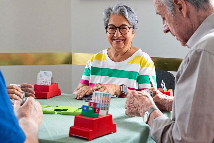 Eine Gruppe älterer Menschen sitzt an einem Tisch und spielt Bridge. Im Vordergrund sind Kartenspiele und rote Kartenhalter zu sehen. Eine Frau mit grauen Haaren, Brille und einem bunt-gestreiften T-Shirt lächelt in die Kamera.