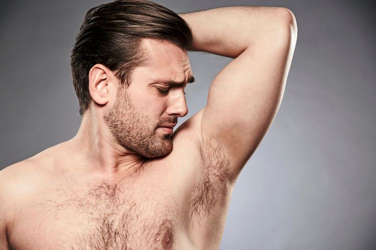 Ein shirtloser Mann riecht an seiner Achsel und schaut ernst, vor grauem Hintergrund.