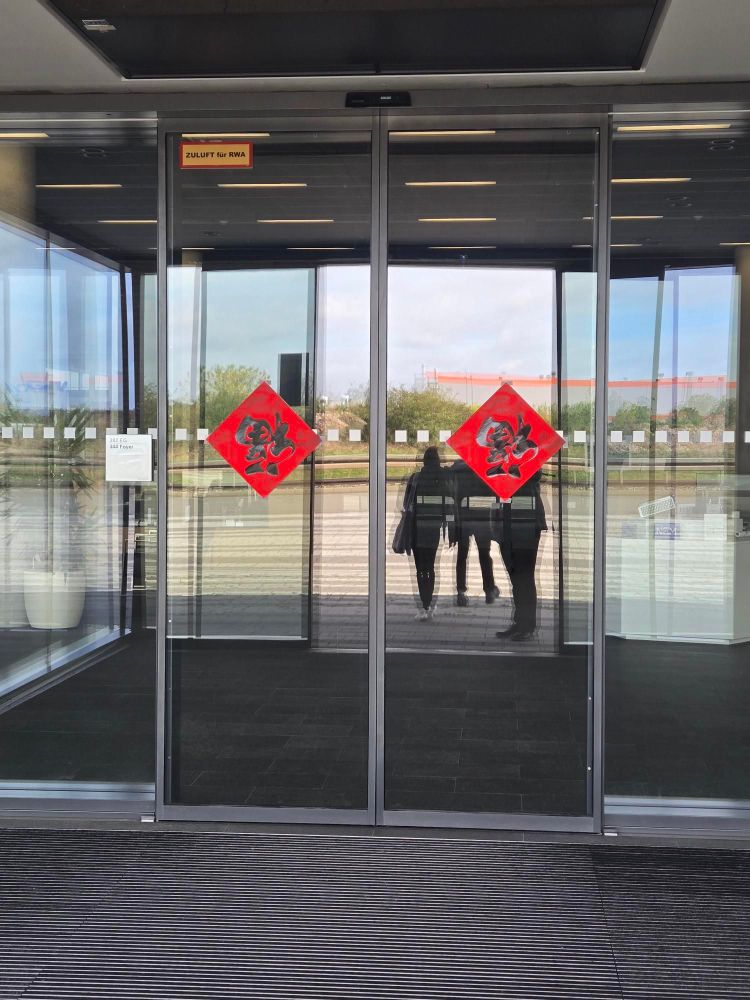 Eine automatische Tür; darauf ein chinesisches Zeichen in rot; im Glas der Tür spiegeln sich Personen