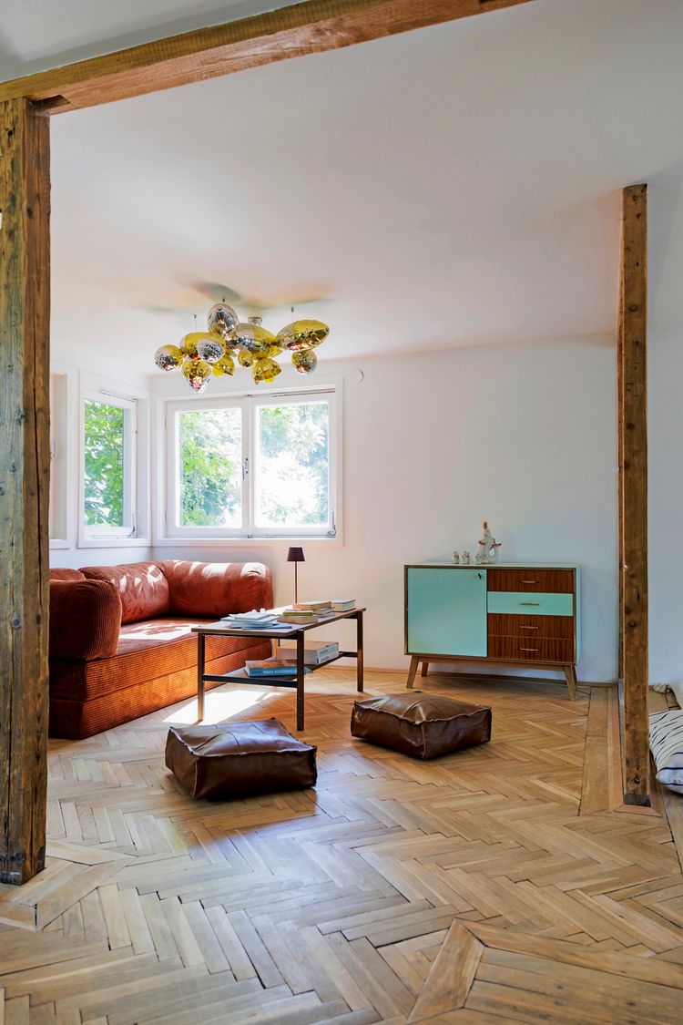 Wohnzimmer mit Holzboden und sichtbaren Holzbalken. Eine orangefarbene Samtcouch steht vor einem Fenster mit Blick ins Grüne. Ein Couchtisch mit Büchern und eine blau-braune Kommode befinden sich im Raum. Zwei braune Sitzkissen liegen auf dem Boden, und eine auffällige, moderne Deckenlampe hängt von der Decke.