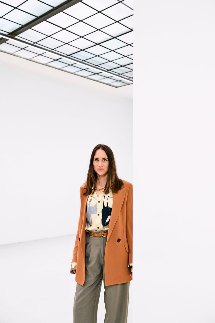 Eine Frau steht in einem hellen Raum mit einem gläsernen, gitterartigen Dach. Sie trägt einen orangefarbenen Blazer, eine gemusterte Bluse und beigefarbene weite Hosen. Sie schaut direkt in die Kamera.