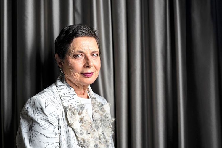 Eine Frau mit kurzen dunklen Haaren posiert vor einem dunklen Vorhang. Sie trägt ein helles Oberteil mit hellgrauen Mustern und dekorativen Details.