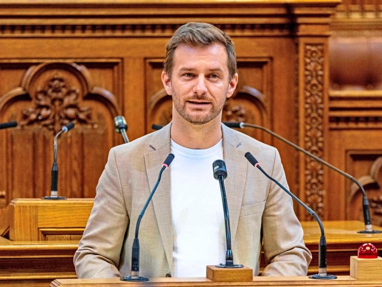 Ein Mann steht an einem Rednerpult im Wiener Rathaus und spricht in ein Mikrofon. Im Hintergrund ist die detailreiche Holzvertäfelung des Saals zu sehen.