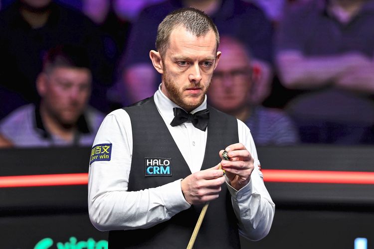 Ein Snookerspieler in weißem Hemd, schwarzer Weste und Fliege steht am Tisch und präpariert seinen Queue mit Kreide. Im Hintergrund sind unscharf Zuschauer zu sehen.