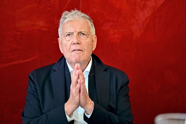 Ein älterer Mann mit grauem Haar in einem dunklen Anzug sitzt vor einem roten Hintergrund. Er hat die Hände vor seinem Gesicht gefaltet und einen nachdenklichen Ausdruck.