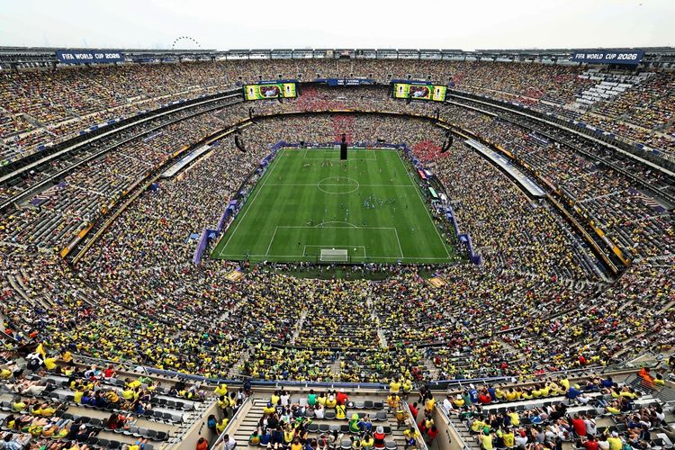 Weitwinkelaufnahme eines vollen Stadions in East Rutherford während der Fußball-Weltmeisterschaft 2026. Spieler wärmen sich auf dem Spielfeld vor einem Achtelfinalspiel zwischen Brasilien und Norwegen auf. Zuschauer füllen die Ränge, viele tragen gelbe Trikots.