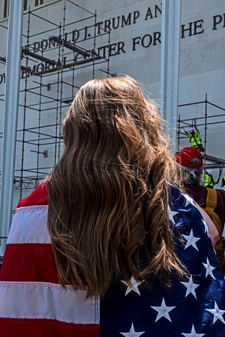 Eine Person mit langen braunen Haaren, die in eine amerikanische Flagge gehüllt ist, steht vor einem Gebäude mit Gerüsten. Im Hintergrund ist eine Fassade mit teilweise lesbarem Text und arbeitende Personen auf dem Gerüst zu sehen.