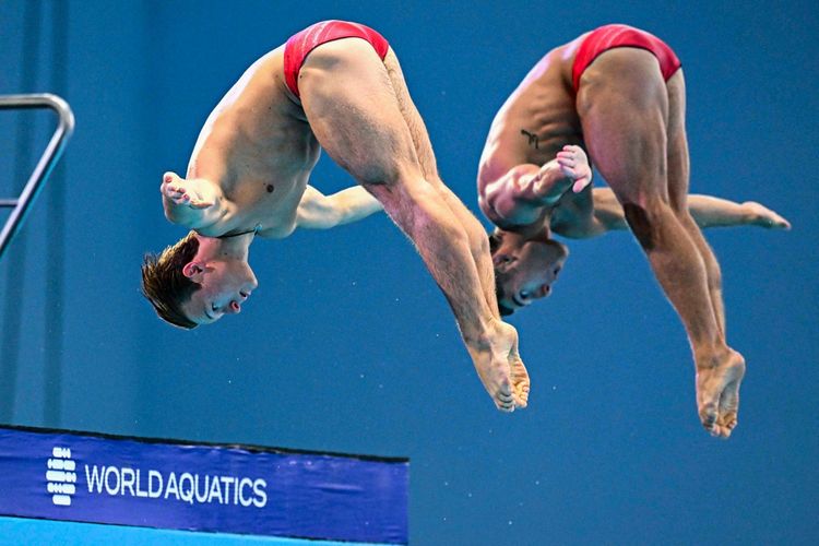 Zwei Wasserspringer in roten Badehosen führen synchron einen Rückwärtssalto vom 10-Meter-Turm während eines Wettkampfs aus. Im Hintergrund der Schriftzug „World Aquatics“.