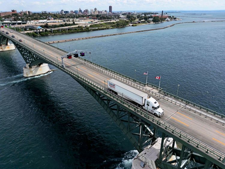 Luftaufnahme der Peace Bridge, die Kanada mit den USA verbindet, über einem breiten Gewässer. Ein weißer Lkw mit der Aufschrift „Capital Southern Transportation, Inc.“ fährt über die Brücke. Im Vordergrund wehen die Flaggen der USA und Kanadas. Im Hintergrund sind eine Stadtlandschaft und Ufergebiete zu sehen.