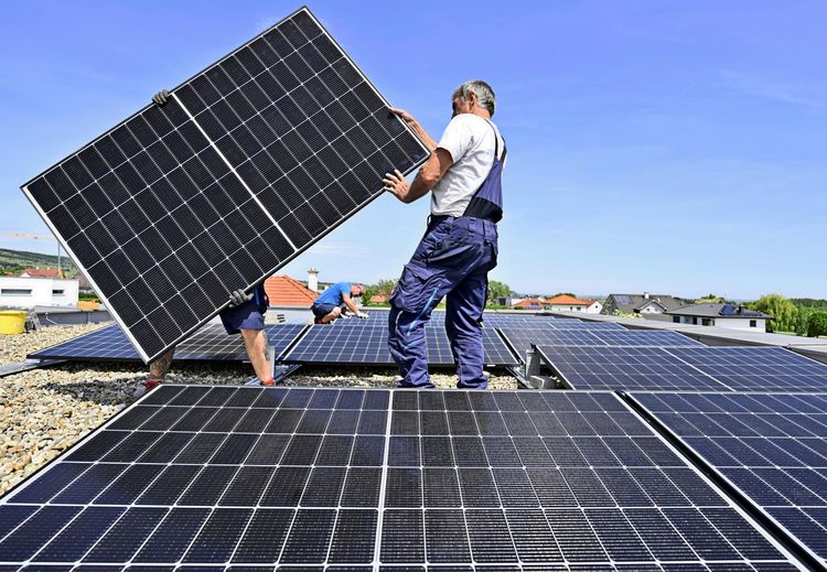 Arbeiter montieren Photovoltaikmodule auf einem Flachdach bei sonnigem Wetter in Purbach am Neusiedler See. Im Hintergrund sind Wohnhäuser und blauer Himmel zu sehen.