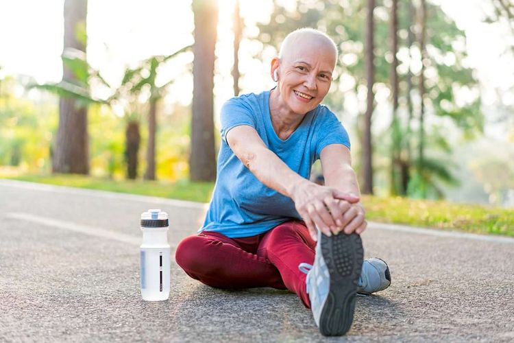 Frau, die offensichtlich an Krebs erkrankt ist (Glatze), sitzt in Sportkleidung auf dem Boden und dehnt nach dem Laufen.