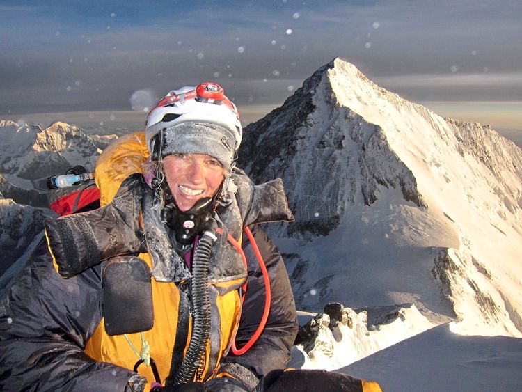 Eine Person in Bergsteigerkleidung, inklusive Helm und Sauerstoffmaske, lächelt in die Kamera. Im Hintergrund sind schneebedeckte, majestätische Berge unter dramatischem, goldenem Licht zu sehen.