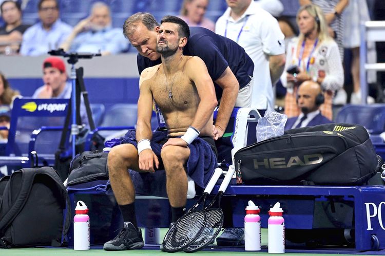 Ein Tennisspieler sitzt mit freiem Oberkörper auf einer Bank, während ein Physiotherapeut seinen Rücken behandelt. Neben ihm liegen Tennistaschen und Trinkflaschen. Im Hintergrund sind Zuschauer und Offizielle zu sehen.