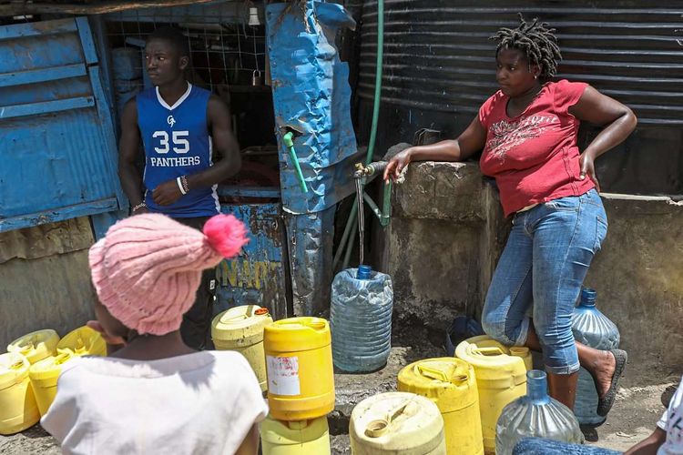 Zwei Frauen stehen vor einem Wasserhahn, umgeben von zahlreichen Kunststoffkanistern.