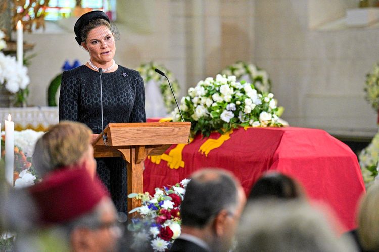 Eine Frau in schwarzer Trauerkleidung und schwarzem Hut mit Schleier spricht am Rednerpult während eines Trauergottesdienstes. Im Hintergrund befindet sich ein Sarg mit rotem Tuch und Blumenschmuck.