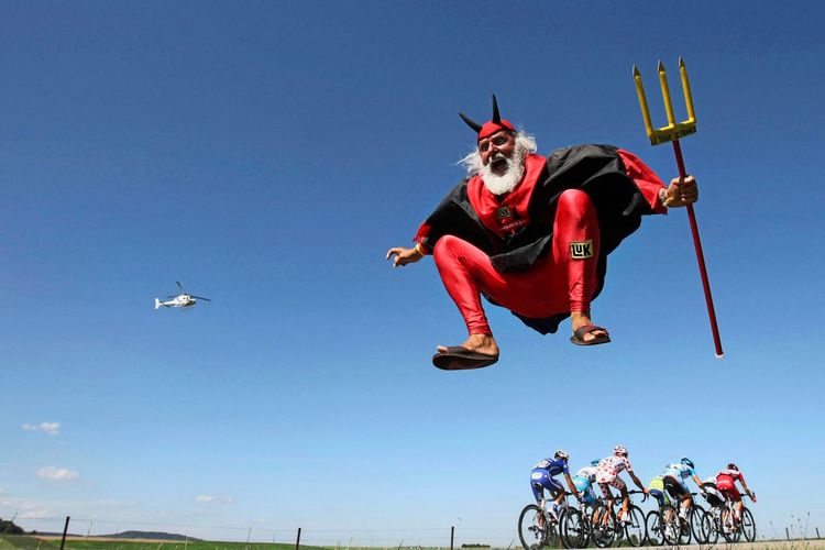 Ein Mann in einem auffälligen roten Teufelskostüm mit Hörnern, Umhang und Dreizack springt in die Luft. Im Hintergrund fahren Radrennfahrer in einer Gruppe während einer Etappe der Tour de France und ein Hubschrauber fliegt am Himmel.