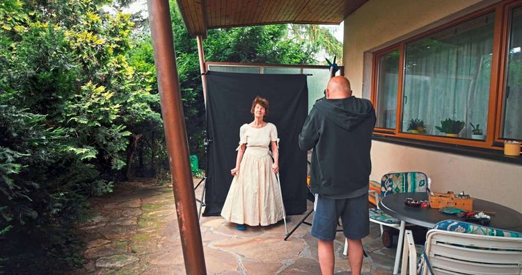 Zu sehen ist eine Fotograf, der im Außenbereich eines Einfamilienhauses eine Frau im weißen Kleid, seine Mutter, fotografiert.