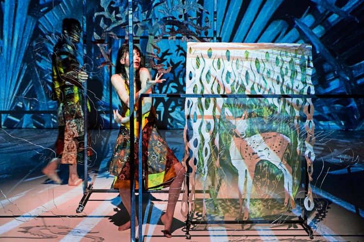 Eine Frau steht in einer künstlerischen Bühneninstallation mit Projektionen von Pflanzen, Mustern und einem Hirsch. Glaswände, durchscheinende Stoffe und Lichteffekte erzeugen eine surreale Atmosphäre.