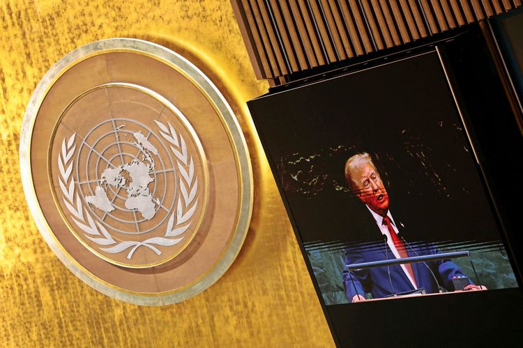 Ein großer Saal mit dem Emblem der Vereinten Nationen (eine Weltkarte, umgeben von Lorbeerzweigen) an einer goldfarbenen Wand. Rechts im Bild zeigt ein Bildschirm eine Person am Rednerpult.
