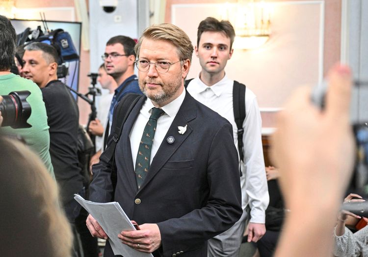 Ein Mann mit Brille, Anzug und Krawatte, der Dokumente in der Hand hält, betritt einen Raum. Im Hintergrund sind weitere Personen, darunter eine mit einem weißen Hemd und Rucksack, sowie Kamerateams zu sehen. Die Szene scheint Teil einer offiziellen Anhörung oder Veranstaltung zu sein.