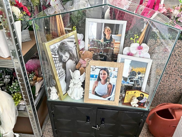 Ein gläserner Schaukasten mit mehreren gerahmten Fotos von Frauen, darunter ein Schwarz-Weiß-Bild und farbige Porträts. Im Kasten befinden sich außerdem Dekorationsgegenstände wie weiße Engelsfiguren, eine Orchideenblüte und ein Plüschtier. Im Hintergrund sind weitere Blumenarrangements zu sehen. Der Schaukasten steht auf einem schwarzen Schrank, daneben steht eine rote Gießkanne.