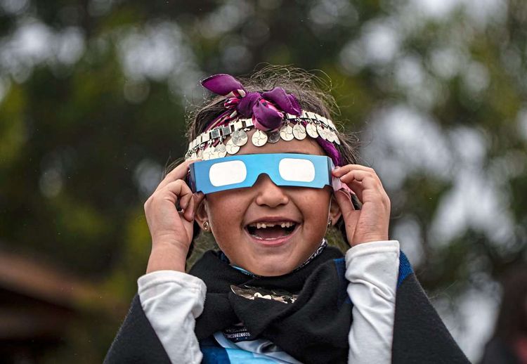 Junges Mädchen mit Haarschmuck hält sich eine Schutzbrille für die Sonnenfinsternis vor die Augen.