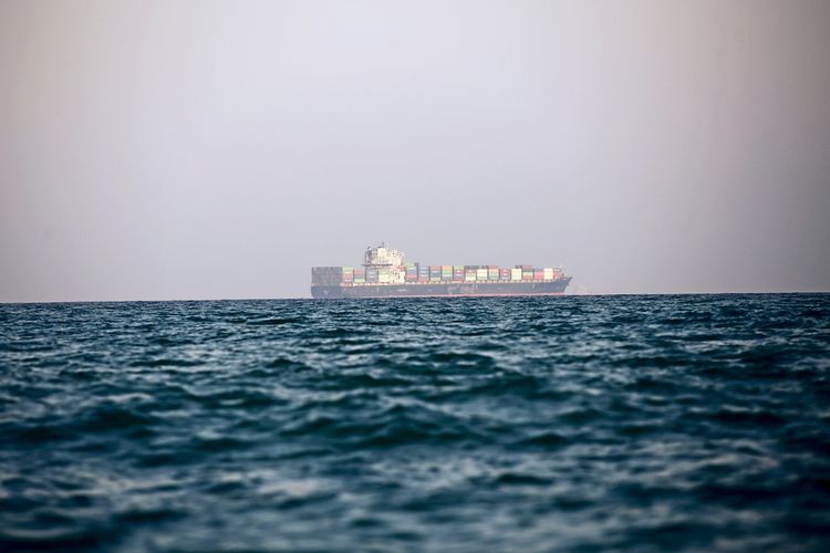 Ein Frachtschiff ist in der Ferne zu sehen, vor einem Hintergrund aus ruhiger, blauer See und grauem Himmel. Das Schiff transportiert bunte Container und ist vor der Küste zwischen den iranischen Inseln Qeshm und Larak im Persischen Golf verankert.