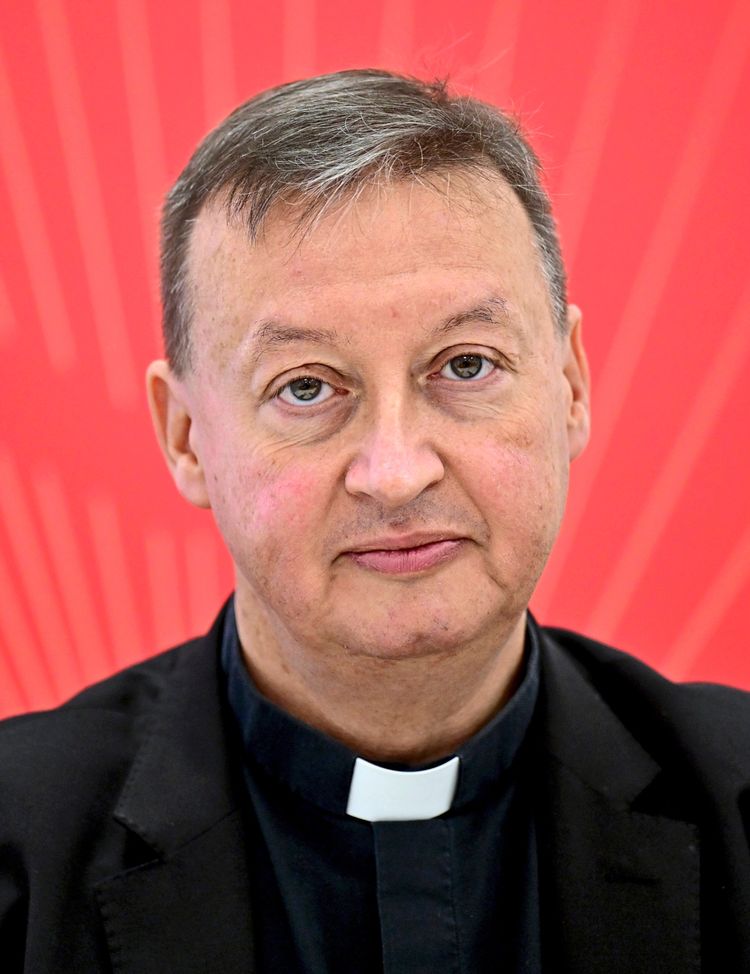 Ein Mann in priesterlicher Kleidung vor einem roten Hintergrund während einer Pressekonferenz.