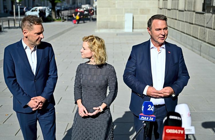 Drei Personen stehen im Freien vor Mikrofonen und geben ein Statement ab. Links ein Mann im dunklen Anzug, in der Mitte eine Frau in einem grauen Kleid, und rechts ein Mann im dunklen Anzug mit weißem Hemd. Im Hintergrund ein Straßenbereich mit Gebäuden.