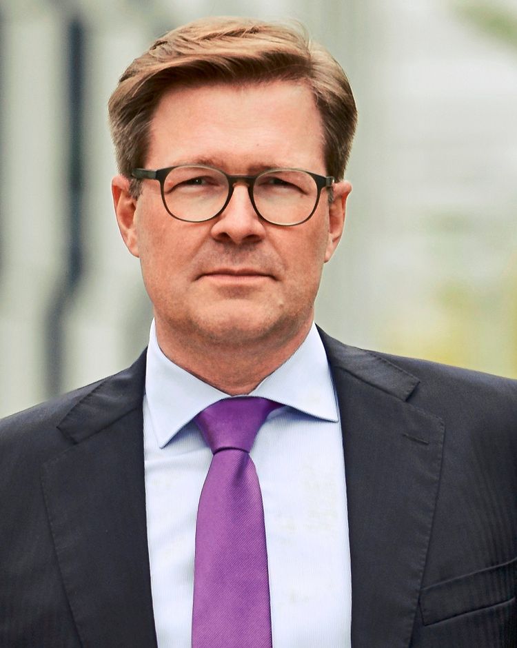 Ein Mann in einem dunklen Anzug, hellem Hemd und lilafarbener Krawatte, im Freien vor einem unscharfen Hintergrund.