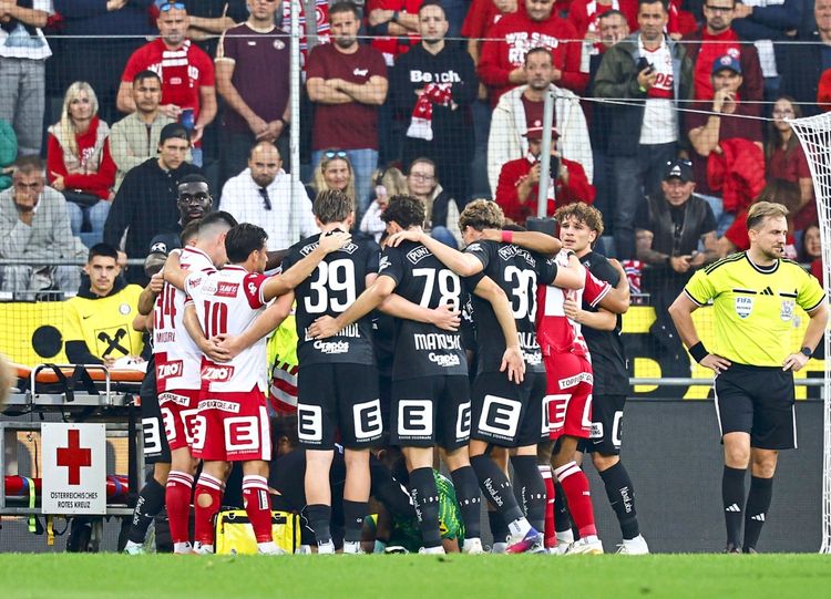 Spieler von GAK und Sturm Graz bilden einen Kreis um den am Boden liegenden, verletzten Daniil Khudyakov (Sturm Graz), während eines Spiels der Admiral Bundesliga (9. Runde) im Stadion Graz-Liebenau, am 18. Oktober 2026. Ein Schiedsrichter steht daneben, während Zuschauer im Hintergrund zu sehen sind. Ein Sanitätskoffer des Österreichischen Roten Kreuzes ist im Vordergrund sichtbar.