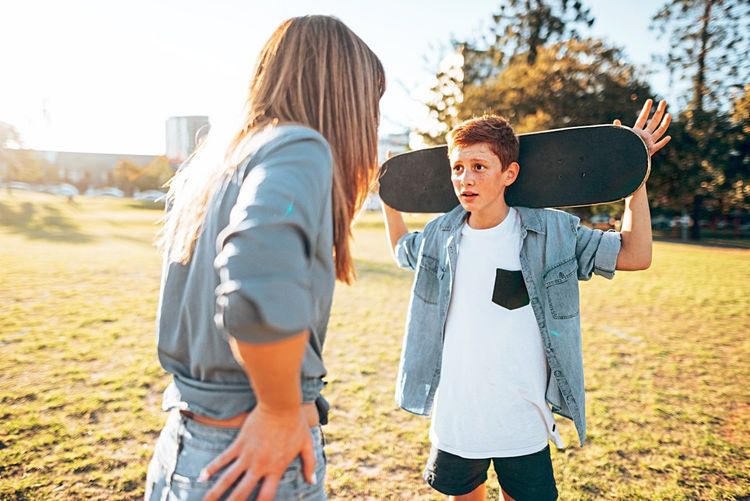 Mutter und jugendlicher Sohn stehen auf einer Wiese und streiten sich, er hält ein Skateboard hinter dem Kopf