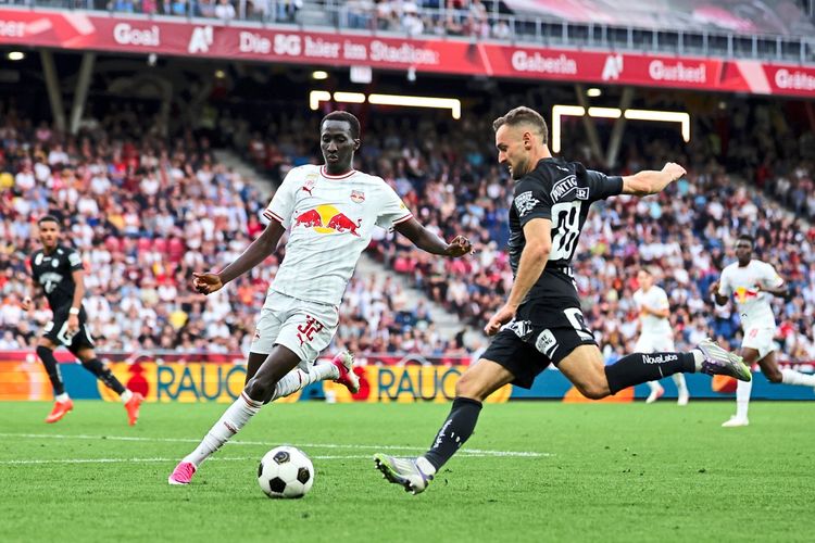 Ein Fußballspiel der Admiral Bundesliga zwischen Red Bull Salzburg und SK Sturm Graz. Im Vordergrund stehen zwei Spieler im Zweikampf um den Ball: ein Spieler von Salzburg in einem weißen Trikot und ein Spieler von Sturm Graz in einem schwarzen Trikot. Im Hintergrund sind Zuschauer und weitere Spieler erkennbar.